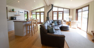 Open-plan living area with a leather sectional sofa facing large glass doors; sunlight fills the space, wood floors, and teal cushions add color.