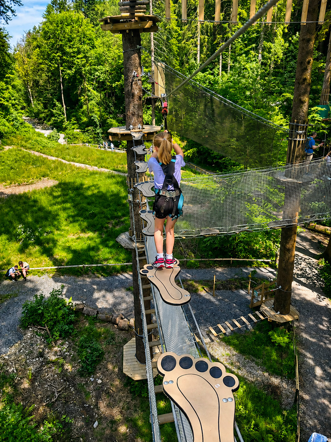 Footbridge Obstacle at Go Ape Grizedale Forest - Lakes Lodges Young girl in a harness crosses a high ropes course on footprint-shaped stepping planks among tall trees in a forest park.